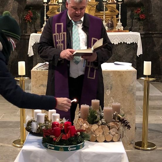 Ein Priester liest ein Buch am Altar mit mehreren Kerzen, während eine andere Person eine Kerze anzündet.