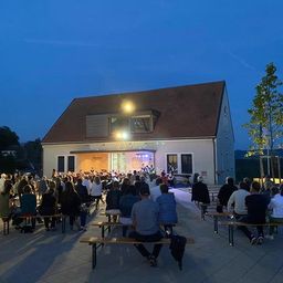 Ein Abendtreffen mit einer Band, die vor einem Gebäude spielt, Menschen auf Bänken sitzen und Fahnen auf Stangen.