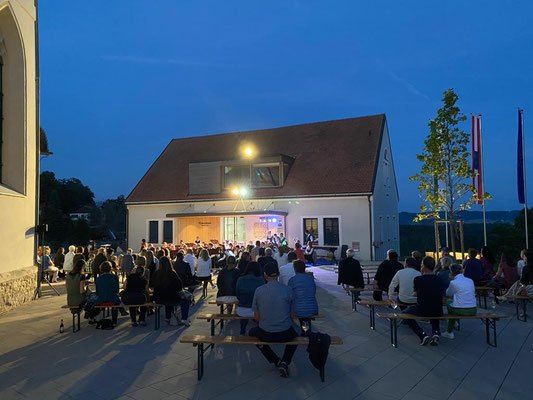 Ein Abendtreffen mit einer Band, die vor einem Gebäude spielt, Menschen auf Bänken sitzen und Fahnen auf Stangen.