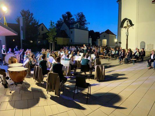 Ein Blasorchester spielt bei Dämmerung vor einer Kirche im Freien, Musiker sitzen auf Stühlen und ein Publikum schaut zu.
