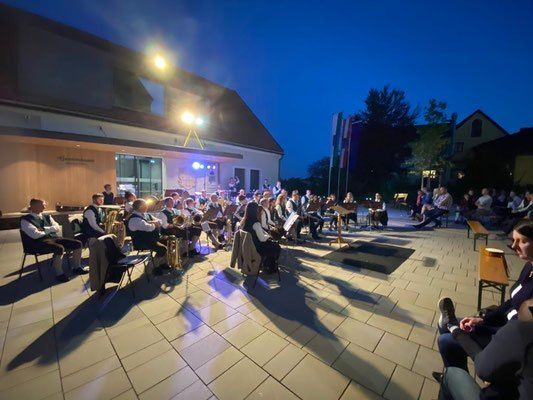 Eine Band spielt auf einer gefliesten Freiluftbühne bei Nacht, mit vielen Menschen in Stühlen um sie herum und einem Gebäude im Hintergrund.