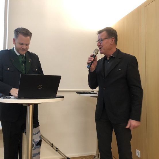 Zwei Männer stehen neben einem Tisch mit einem Laptop. Einer hält ein Mikrofon und scheint zu sprechen. Eine Holzwand ist hinter ihnen.