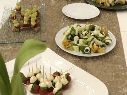 Ein Tisch ist mit verschiedenen Platten von Vorspeisen gedeckt, darunter eine mit einer Mischung aus Gemüse und Oliven, eine andere mit einem Gemüsesalat und eine dritte mit kleinen Häppchen auf Spießen. Ein grünes Blatt fügt einen Farbakzent hinzu.