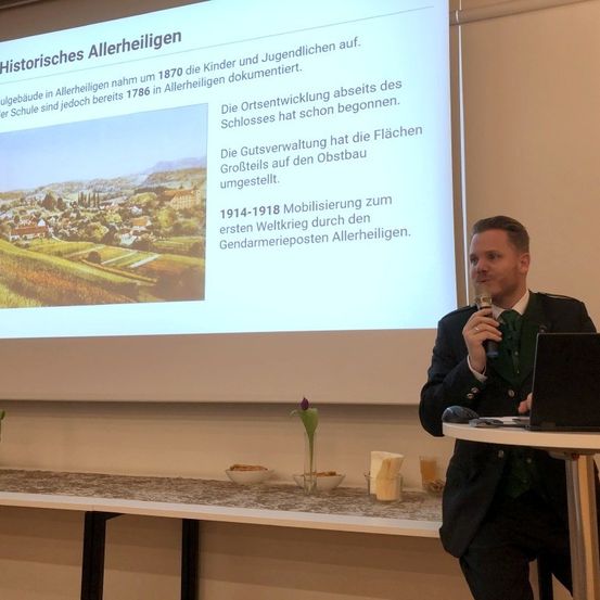 Ein Mann steht vor einer Projektionsleinwand und hält eine Präsentation. Die Leinwand zeigt Text über historische Allerheiligen.