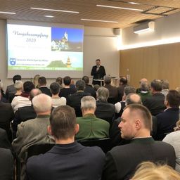 Ein Mann hält eine Rede vor einer Projektionsleinwand, die ein Bild einer Kirche mit dem Text 'Neujahrsempfnag 2020' zeigt. Das Publikum sitzt aufmerksam in Stühlen.