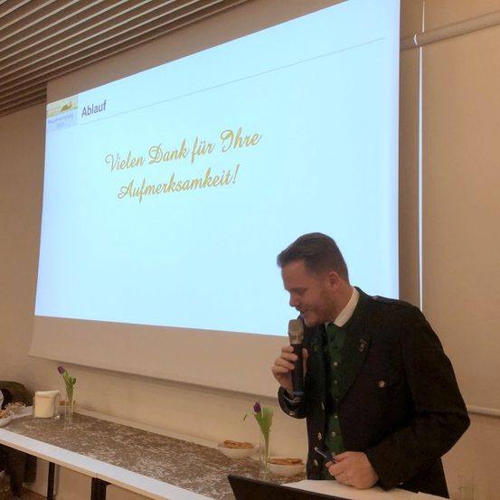 Ein Mann spricht in ein Mikrofon vor einer Leinwand, die 'Vielen Dank für Ihre Aufmerksamkeit!' zeigt. Ein Tisch mit Blumen und Essen steht in der Nähe.