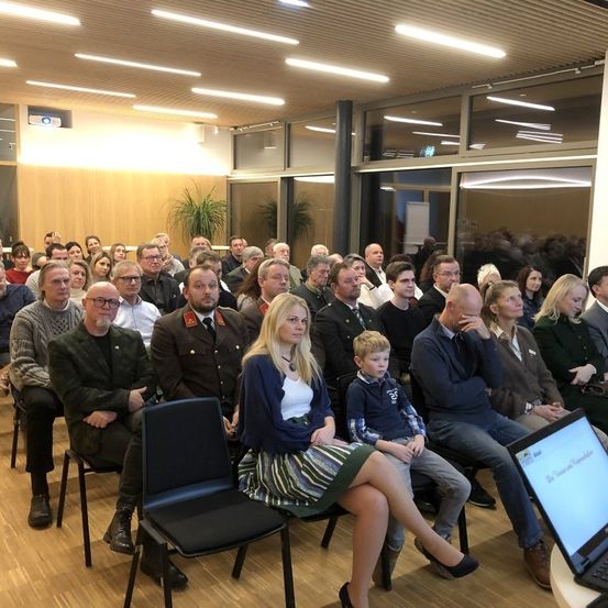 Eine große Gruppe von Menschen sitzt in einem Konferenzraum, einige tragen Brillen und Uniformen. Ein Laptop-Bildschirm ist rechts sichtbar, und große Fenster und Deckenleuchten sind im Hintergrund.
