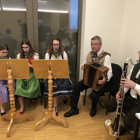 Fünf Musiker, drei Frauen und zwei Männer, spielen Instrumente. Ein Mann spielt Akkordeon, ein anderer Saxophon und die Frauen Klarinetten. Sie sitzen in einem Raum mit Holzboden.