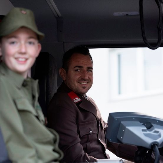 Zwei Personen sitzen in einem Fahrzeug, einer trägt eine grüne Militäruniform und der andere eine braune Uniform mit einem roten Abzeichen auf der Schulter. Beide lächeln und schauen nach vorne.