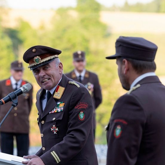 Ein uniformierter Offizier hält eine Rede an einem Rednerpult, während zwei weitere uniformierte Offiziere hinter ihm stehen, ein Mikrofon davor.