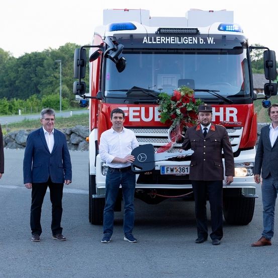 Fünf Männer stehen vor einem roten Feuerwehrauto. Ein Mann hält einen Blumenstrauß und einen Schlüssel. Ein anderer Mann hält eine Plakette.
