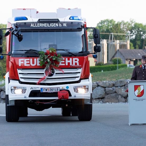 Ein Feuerwehrwagen mit den Worten 'ALLERHEILIGIEN b. W.' und 'FEUERWAHR' auf der Vorderseite ist geparkt. Eine Person steht an einem Rednerpult mit Mikrofon.