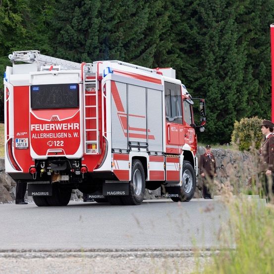 Ein roter Feuerwehrwagen ist am Straßenrand geparkt, neben ihm stehen zwei Feuerwehrleute in Uniform. Der Wagen ist mit der Nummer '122' und der Aufschrift 'Feuerwehr' gekennzeichnet und mit einer Leiter ausgestattet. Hinter dem Wagen steht eine Fahne.