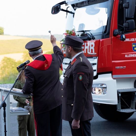 Zwei Männer in Uniformen stehen neben einem roten Feuerwehrauto. Einer hebt die Hand, wahrscheinlich bei einer Rede. Eine Frau hält ein Buch und ein Mikrofon hinter ihnen.