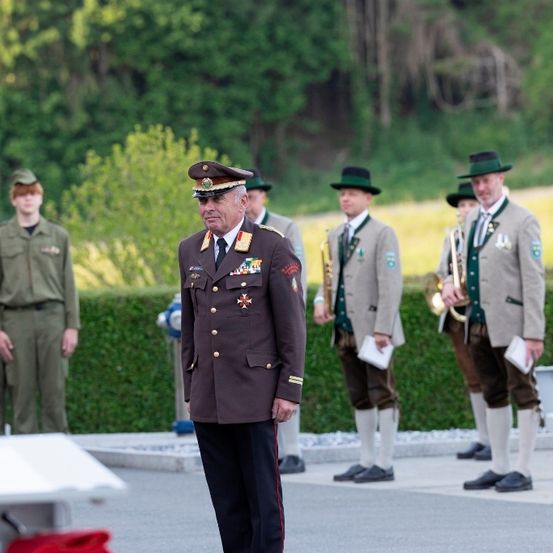 Eine formelle Zeremonie mit Militärangehörigen, einschließlich eines hochrangigen Offiziers in traditionellen Uniformen, steht im Freien mit Musikinstrumenten und Grün im Hintergrund.