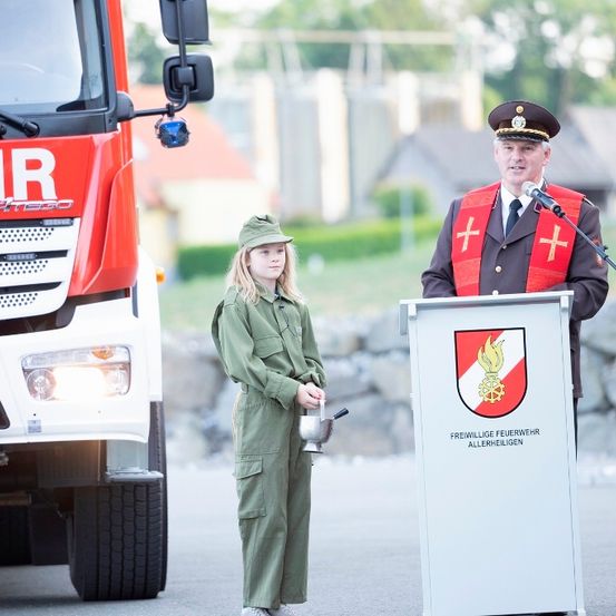 Ein Feuerwehrmann in Uniform spricht eine junge Frau in grüner Kleidung vor einem Feuerwehrauto an.