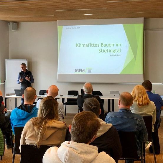Eine Innenpräsentation mit einem Mann, der an einer Leinwand mit dem Titel 'Klimafittes Bauen im Stiefingtal' präsentiert. Mehrere Personen sitzen vor ihm in einem Klassenzimmer.