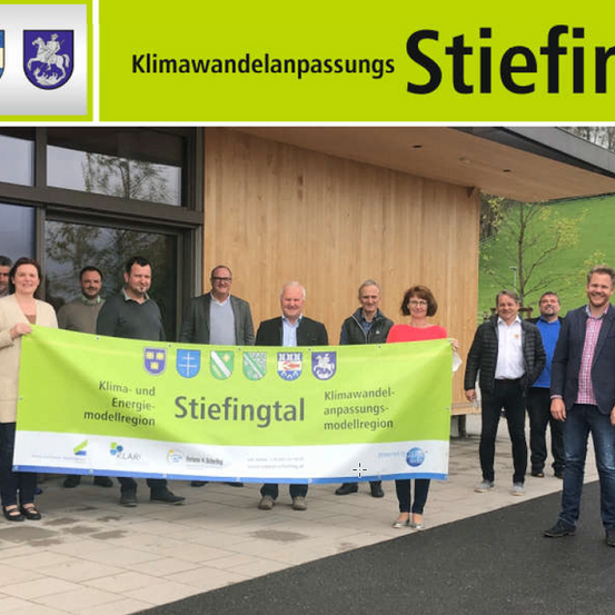 Eine Gruppe von Menschen steht vor einem Gebäude und hält ein Banner mit verschiedenen Logos. Sie lächeln für ein Foto.