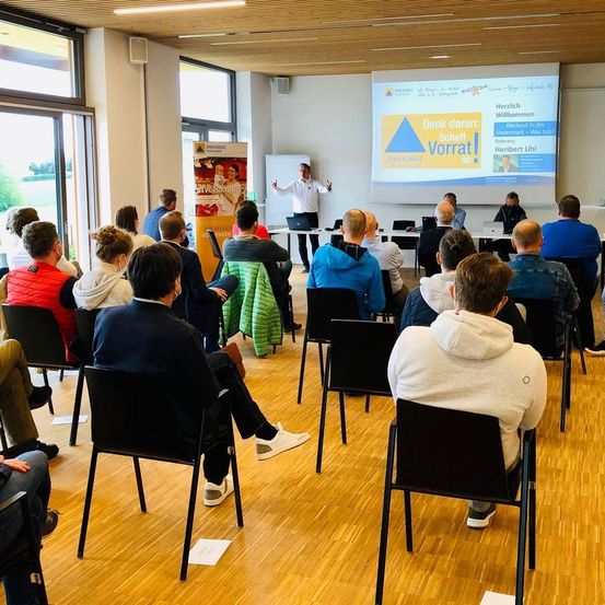 Ein Raum mit Holzboden und Glasfenstern hat eine Projektionsfläche. Mehrere Personen sitzen auf Stühlen und ein Mann steht vor der Leinwand.