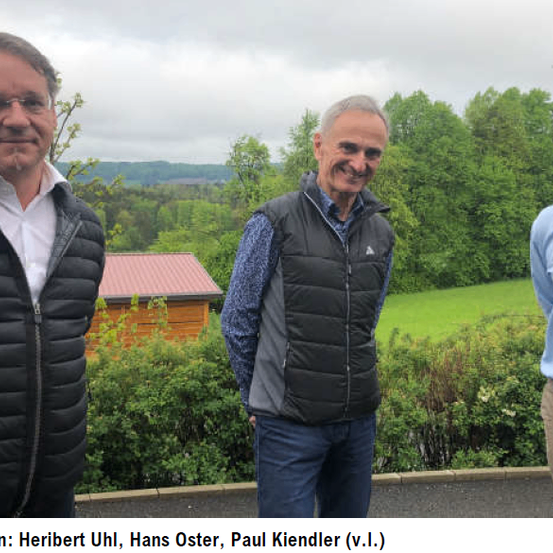 Drei Männer stehen draußen in der Nähe eines kleinen Hauses, lächeln und posieren für ein Foto. Dahinter befindet sich ein Rasenfeld mit Bäumen.