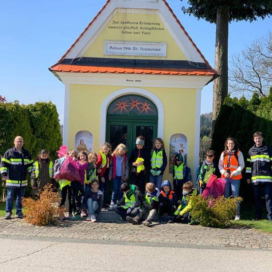 Eine Gruppe von Menschen, wahrscheinlich Freiwillige, steht vor einer kleinen Kapelle. Sie tragen reflektierende Westen und halten Plastiktüten. Die Kapelle hat eine deutsche Inschrift.
