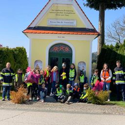 Eine Gruppe von Menschen, wahrscheinlich Freiwillige, steht vor einer kleinen Kapelle. Sie tragen reflektierende Westen und halten Plastiktüten. Die Kapelle hat eine deutsche Inschrift.