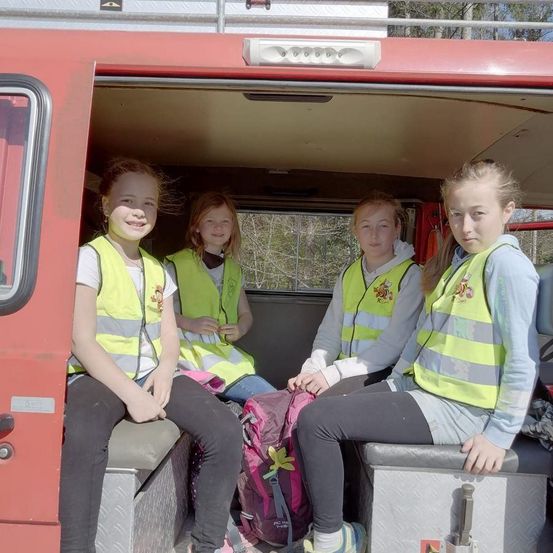Vier Mädchen in Warnwesten sitzen in einem roten Feuerwehrwagen und lächeln für ein Foto. Das Fahrzeug hat ein weißes Dach und ein Glasfenster auf der linken Seite.