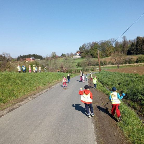 Mehrere Kinder in gelben Westen laufen mit einigen Rechen entlang einer asphaltierten Straße. Sie sind von Grasflächen und Bäumen umgeben.