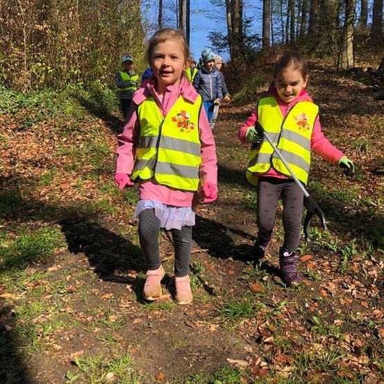 Zwei junge Mädchen in hellen Warnwesten laufen auf einem Waldweg. Sie werden von anderen Kindern begleitet, und eine hält einen Besen. Der Wald ist mit trockenen Blättern und Bäumen gefüllt.