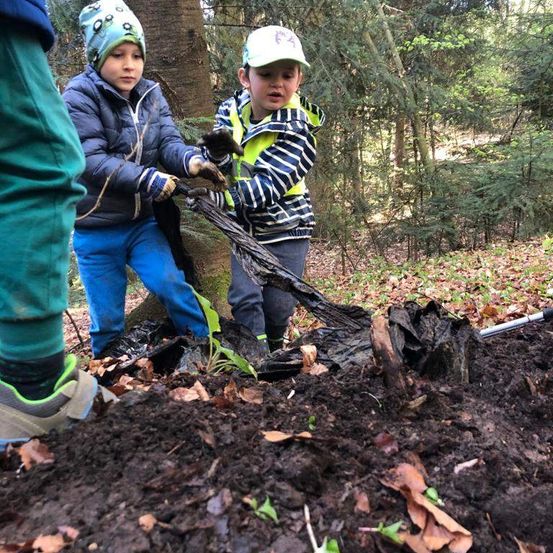 Zwei Kinder pflanzen einen Baum im Wald. Einer hält einen Baumzweig und der andere einen schwarzen Plastiksack. Der Boden ist schlammig mit Blättern.
