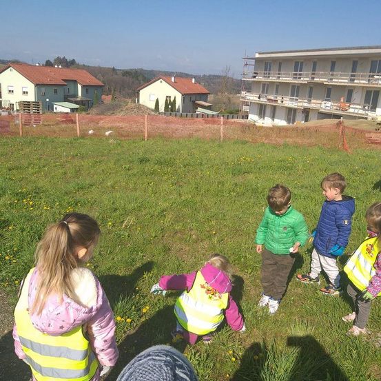 Kinder in Warnwesten stehen auf einem Grasfeld, eines beugt sich, um etwas zu pflücken. Dahinter sind Bauzäune und ein Gebäude im Bau zu sehen. In der Ferne sind Häuser und Hügel zu erkennen.