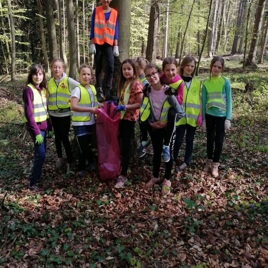 Mehrere junge Mädchen in Warnwesten versammeln sich im Wald. Eine steht auf einem Baum, während andere einen roten Beutel halten. Sie tragen Handschuhe und Schuhe.