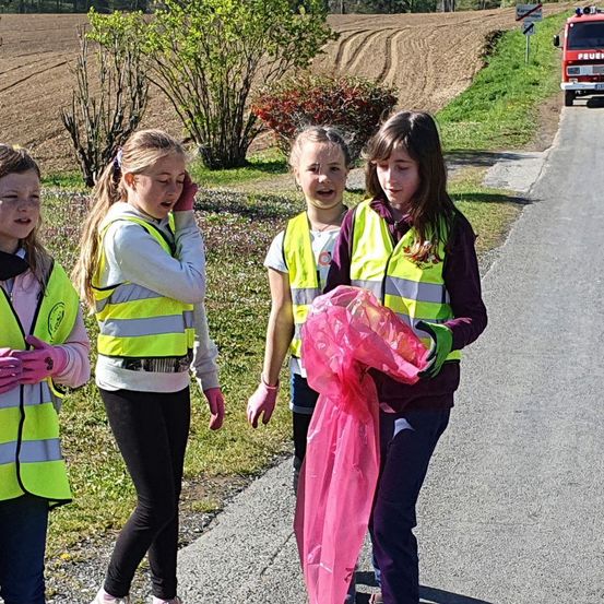 Vier Mädchen in Reflektorwesten gehen am Straßenrand. Eine von ihnen hält einen Plastikbeutel. Dahinter befindet sich ein Feld mit Pflanzen.