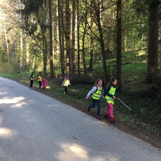 Mehrere Kinder in gelben Westen laufen einen Waldweg entlang, eines hält einen Stock. Sie scheinen an einer Aufräumaktion teilzunehmen.