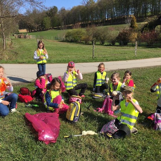 Eine Gruppe von Kindern in gelben Westen sitzt auf dem Gras, einige trinken aus Bechern. Sie sind umgeben von Rucksäcken und Mülltüten, mit einem eingezäunten Feld und Bäumen im Hintergrund.