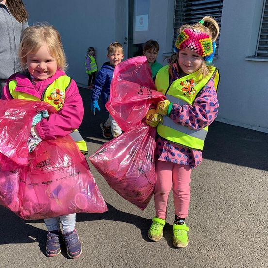 Zwei Mädchen in Sicherheitswesten tragen rosa Taschen und lächeln. Sie tragen bunte Mützen und Turnschuhe. Ein Junge und ein Mädchen in gelben Westen folgen hinter ihnen in der Nähe eines weißen Gebäudes.