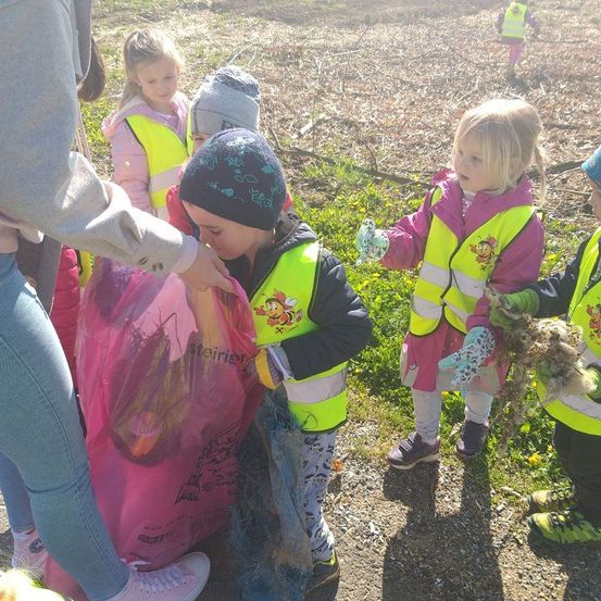 Eine Gruppe von Kindern in Warnwesten sammelt Müll auf einem Feld, wobei eines einen Beutel und andere Pflanzen hält.