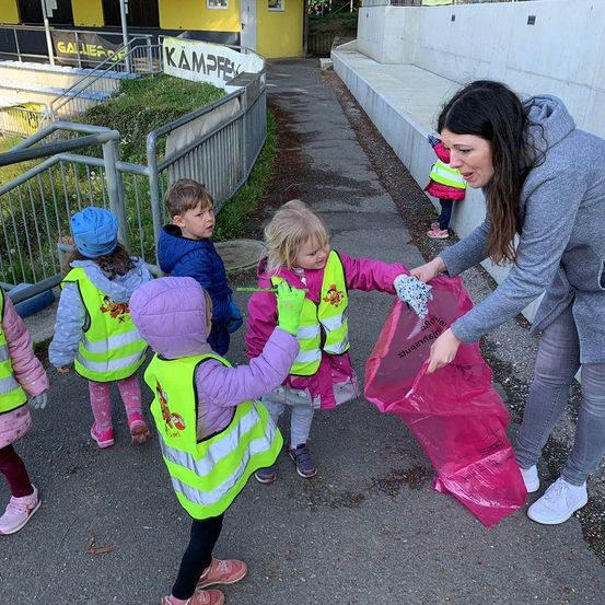 Eine Frau hilft Kindern in neonfarbenen Westen, Müll mit einem rosa Beutel in der Nähe eines gelben Gebäudes zu sammeln.