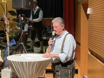 Ein älterer Mann steht an einem Tisch und hält ein Mikrofon. Er scheint eine Rede zu halten. Hinter ihm spielt ein Musiker ein Blechinstrument.