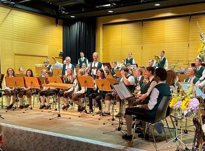 Eine Gruppe Musiker in grüner und weißer traditioneller Kleidung spielt auf einer Bühne mit Musikinstrumenten. Einige sitzen, andere stehen und sie sind von verschiedenen Musikständern und Blumen umgeben.