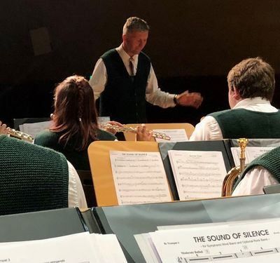 Eine Gruppe von Musikern spielt Instrumente, darunter Flöten und Trompeten, angeleitet von einem Mann in Weste und Krawatte. Vor ihnen liegen geöffnete Notenblätter mit dem Titel 'Der Klang der Stille'.