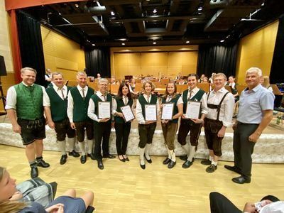 Eine Gruppe von Menschen in grünen Westen und traditioneller Kleidung steht auf der Bühne und hält Urkunden. Sie werden von einigen anderen Personen umgeben, von denen eine sitzt. Die Bühne ist mit Vorhängen und einem Holzboden dekoriert.