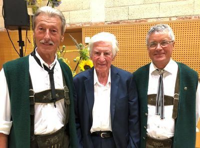 Drei Männer stehen zusammen, alle in traditioneller Kleidung. Der Mann links und rechts trägt grüne Westen mit Krawatten und Trägerhosen, während der Mann in der Mitte einen dunklen Anzug trägt. Hinter ihnen ist eine Wand mit einer Blumenarrangement zu sehen.