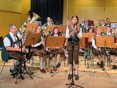 Eine Band tritt auf der Bühne auf, wobei ein Mitglied die Klarinette spielt, während andere verschiedene Instrumente spielen. Das Publikum sitzt im Hintergrund.