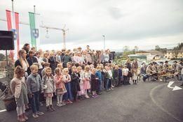 Eine Gruppe von Kindern und Erwachsenen steht in einer Reihe auf einer Straße mit Fahnen und Baugeräten im Hintergrund.