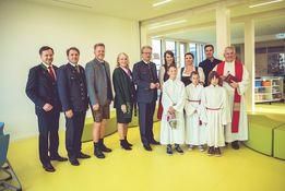 Eine Gruppe von Menschen in formeller Kleidung, einschließlich Kinder in Weiß, steht in einem Raum. Ein Priester in einer weißen Robe ist unter ihnen. Sie posieren für ein Foto.