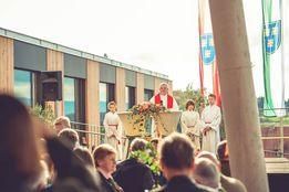 Ein Priester in einer roten Robe steht auf einer Bühne mit einem Mikrofon, umgeben von Menschen in weißen Roben, wahrscheinlich während einer Kirchenveranstaltung.