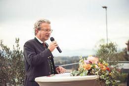Ein älterer Mann in Anzug und Brille spricht bei einer Outdoor-Veranstaltung in ein Mikrofon, mit einem Blumenarrangement und Grünpflanzen im Hintergrund.