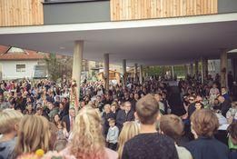 Eine große Menschenmenge versammelt sich unter einem überdachten Bereich. Viele Menschen tragen formelle Kleidung. Einige Personen stehen, andere sitzen. Der Bereich ist von einem modernen Gebäude umgeben.