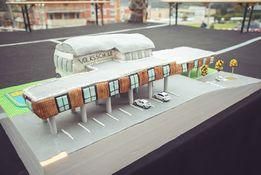 Ein detailliertes Modell eines Bahnhofs mit gekrümmtem Dach, geparkten Autos und Straßenschildern. Gebäude und Bäume sind im Hintergrund zu sehen.